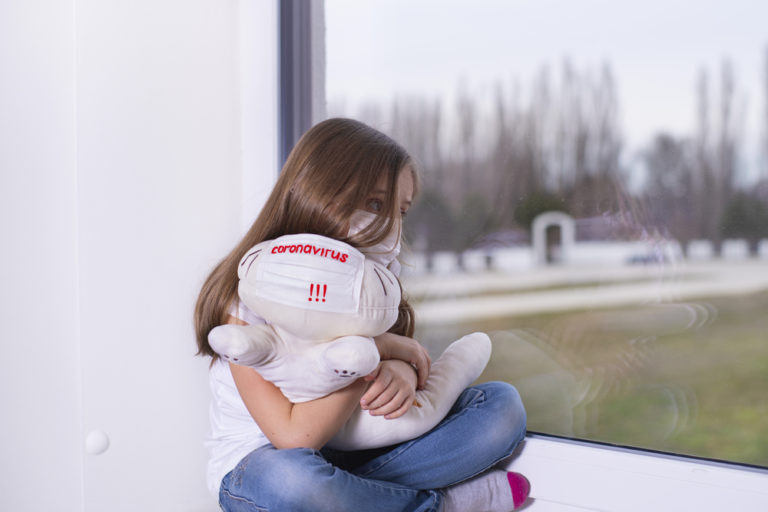 A girl with a toy in a protective mask with the text Coronavirus.A little girl in a white Tshirt with a protective mask from the virus on her face sits by the window with a toy in her hand and is sad